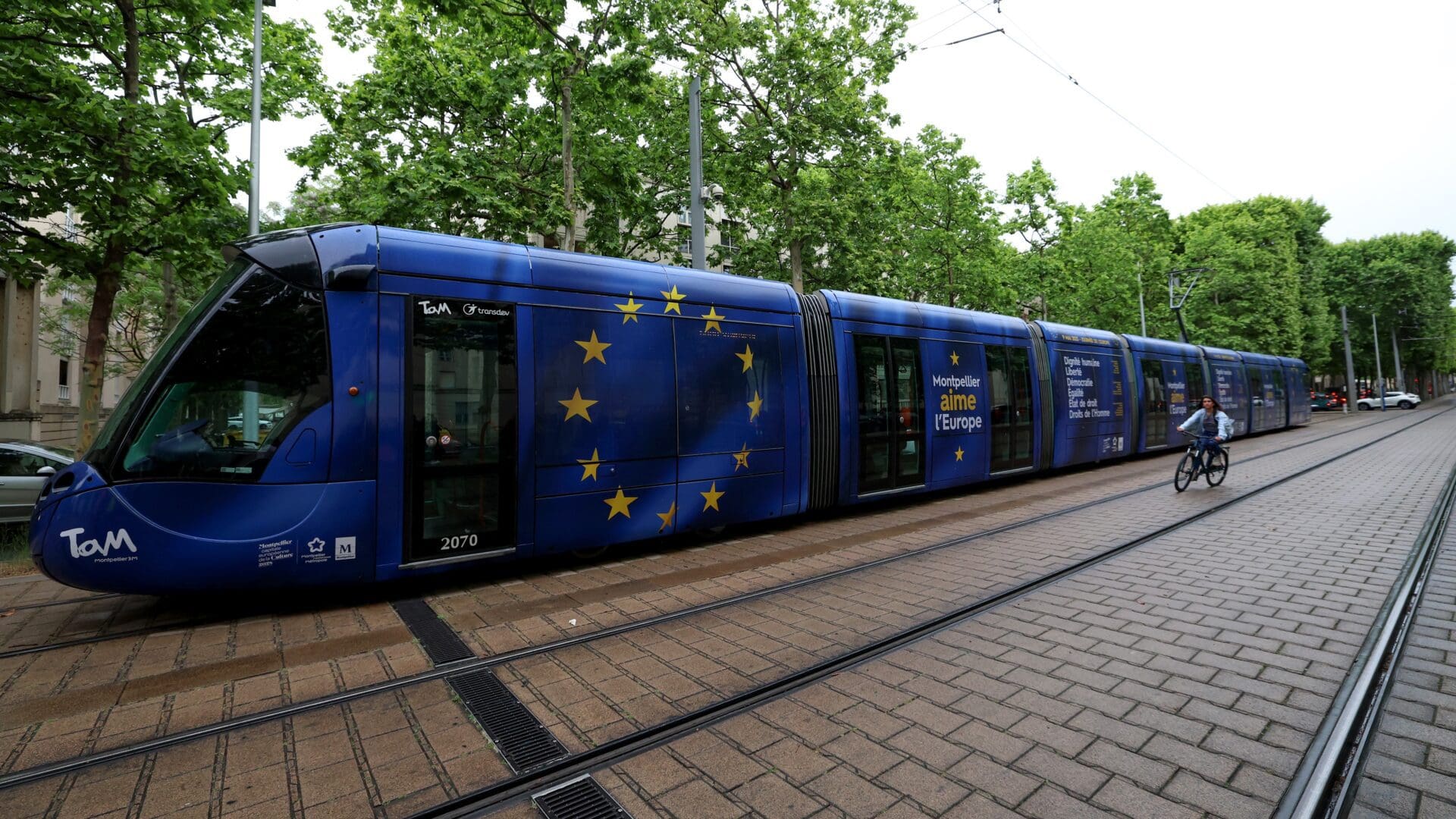 A woman rides a bike next to a tramway, with the colors of European Union, running in the city centre of Montpellier, southern France, on the occasion of Europe Day, on May 9, 2023.