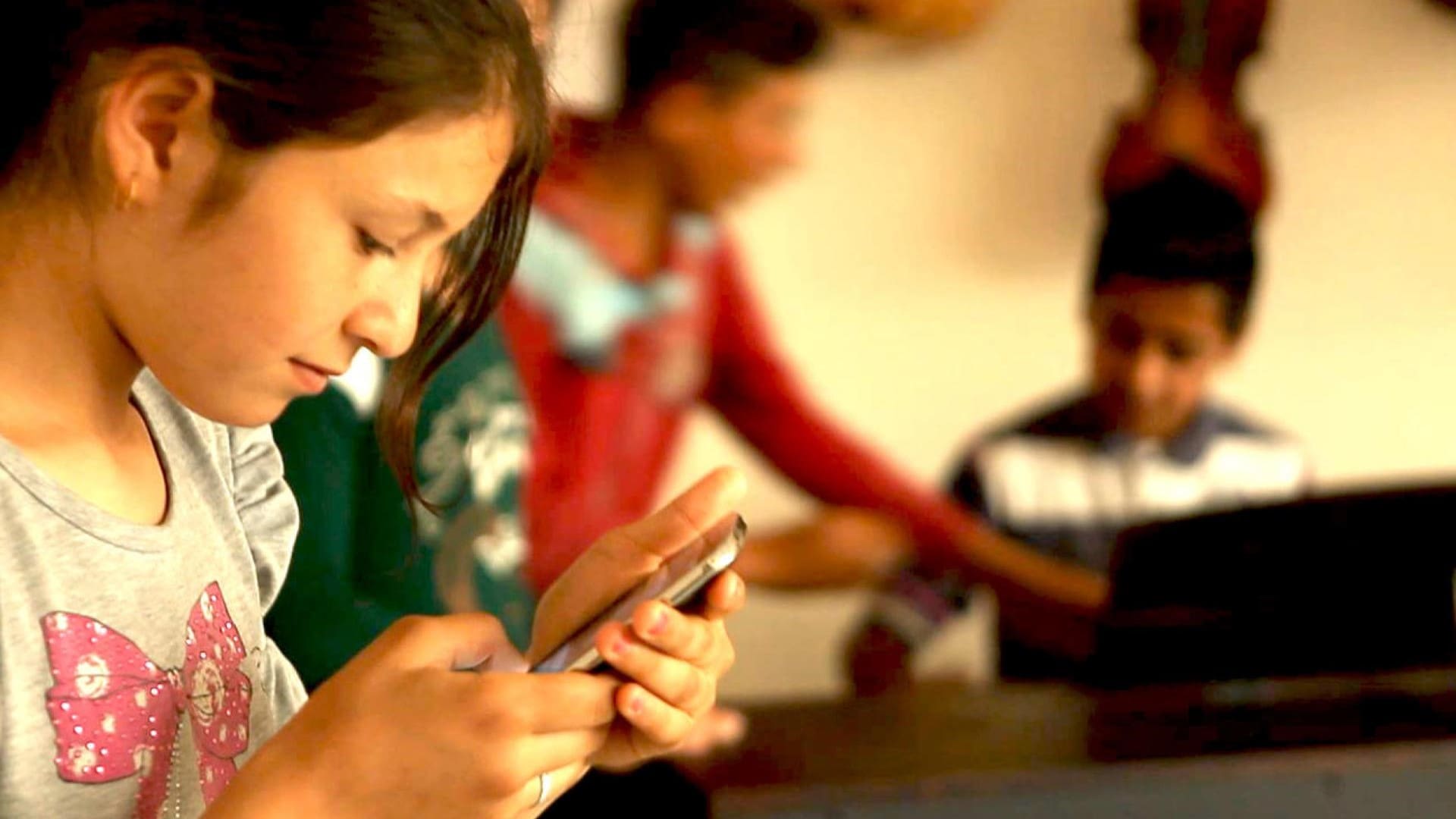 Young girl looking at smartphone with two boys at a piano in the background. (Illustration)