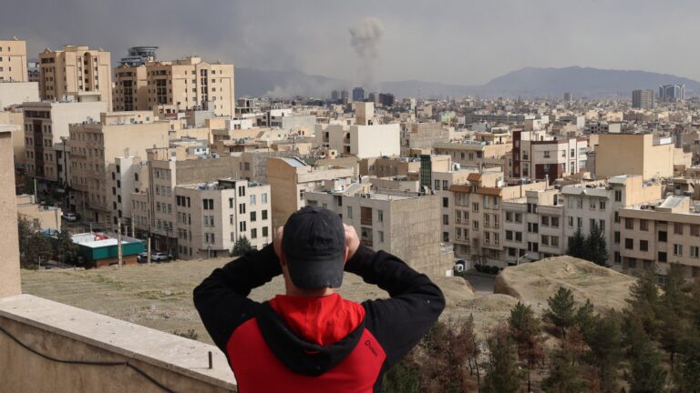 A man looks through biculars as a plume of smoke rises after a military strike on the capital Tehran on March 2, 2026.